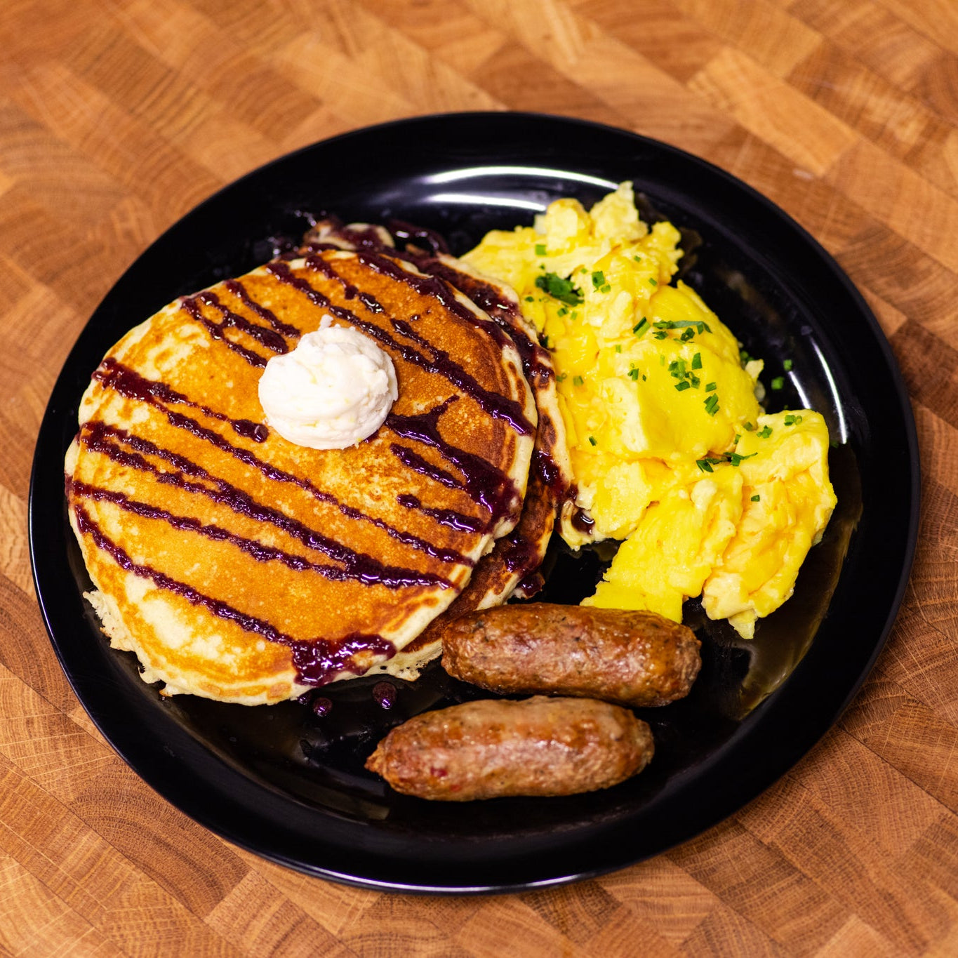 Plate of pancakes with syrup, scrambled eggs, and sausages on a wooden table.
